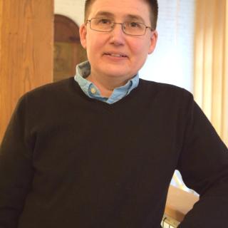 Image of Evin Carvill-Ziemer: white non-binary person with short dark hair and glasses leaning on a railing. Evin is wearing a black sweater over a blue collar shirt.
