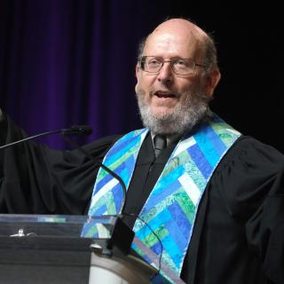 Rev. Chris Buice, arms raised with palms up, wearing a black robe and blue stole while delivering a sermon.