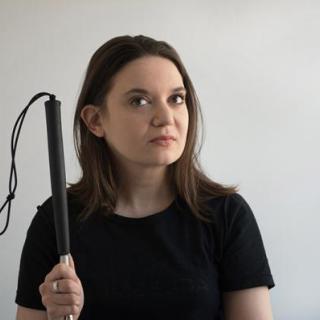 Image of Gretchen Maune: A white non-binary person with dark brown straight hair and blush lipstick, weard a black short sleeve shirt and holding a white cane with a black handle.  