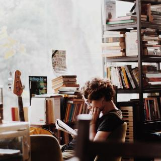 person sits near window reading a book held in their hand with stacks of books on shelves in background