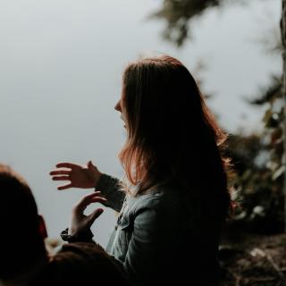 Seen from slightly behind, a person with long hair gestures with their hands while talking.