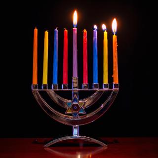 A silver menorah, against a dark background, holds colored candles. The first three candles and the shamash candle are lit.