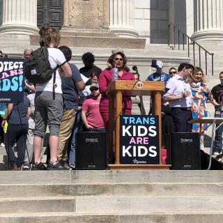 Image from Missouri transgender rights protest rally, with a Black trans woman at a lecture surrounded by supports with a sign that reads "Trans Kids Are Kids - PROMO"