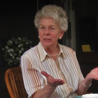 An elderly woman with light skin and silver hair talking with her hands held out in an open gesture.