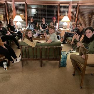A dozen young adult UUs sitting in a circle around a chalice on comfortable furniture in a lounge area surrounded by windows at night. They are smiling and facing the camera.