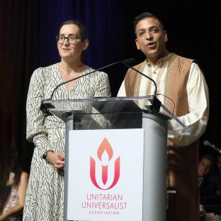 Image of two UU leaders standing at the podium during 2023 General Assembly, Sunday morning worship service. On the left is Rev. Nancy McDonald Ladd, a white woman wearing glasses with dark hair pulled back, wearing a white long-sleeve dress with a white and black pattern. She is looking out to the audience with a grateful expression. On the right is Rev. Manish Mishra-Marzetti, a man of color wearing traditional cream-colored robes and a long vest. Manish is speaking into the microphone looking happy.