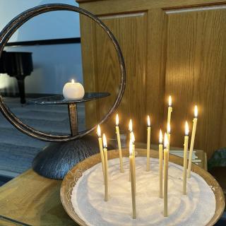 A UU chalice made of bronze metal with large rings and a small white lighted candle in the center. Next to the chalice is a large brown bowl filled with sand with twelve thin white lighted candlesticks. Both the chalice and bowl of candles are on a small wooden table in front of a wooden lectern that is partially visible on the altar of the congregation, with a black grand piano. 