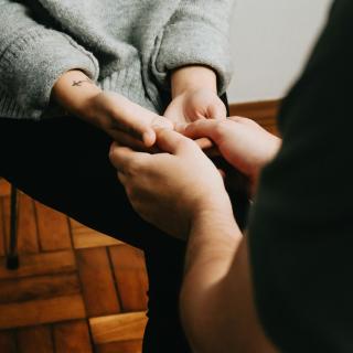 A cropped photo of a person with a wrist tattoo with upturned hands lightly holding the hands of another person, sitting knee-to-knee with them.