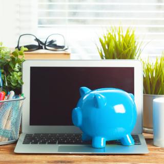Blue piggy bank resting on a laptop, with container of pens to the left and coffee mug on the right. Small plants in the back