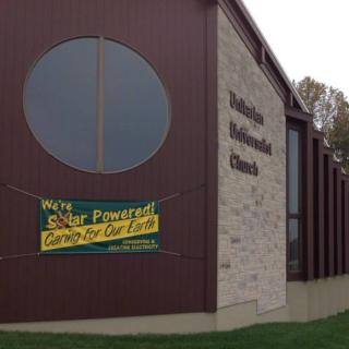 Modern brick and brown wood church building with a large round window and a banner saying "We're solar powered!"