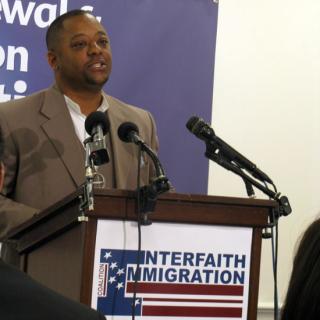 Rev. John Crestwell, speaking at a podium with an 'Interfaith Immigration' sign.