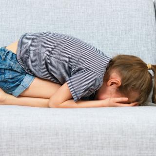 A young child curled up on a couch, her head buried in her hands