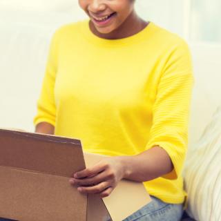 A Black woman, sitting on a couch, smiles as she opens a box. 