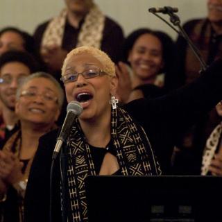As choir members clap in the background, a choir member sings into a microphone 