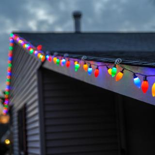 Against a darkening sky, the roofline of a house is strung with colored Christmas lights.