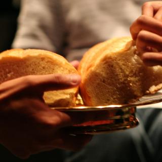 One set of hands hold a plate with a loaf of bread, while another set of hands breaks a piece off for communion.