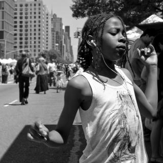 In a black and white photo, a young Black girl dances on a busy city street while listening to music on headphones