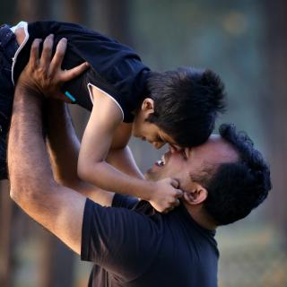 A father and son nuzzle each other; the father looking up, holding his son aloft.