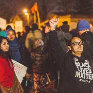 In front of a nighttime crowd, holding signs, a woman in a Black Lives Matter sweatshirt raises her fist into the air.