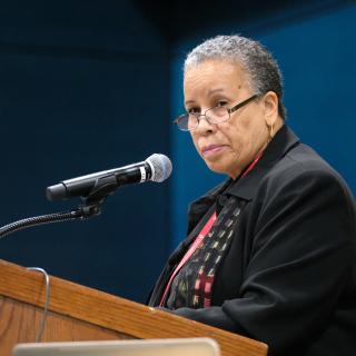 A senior African American looks sternly at the audience from behind a speaker's podium