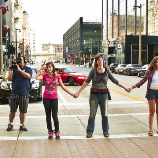 a line of people, of all ages, facing the camera and holding hands, across the intersection of an empty city street