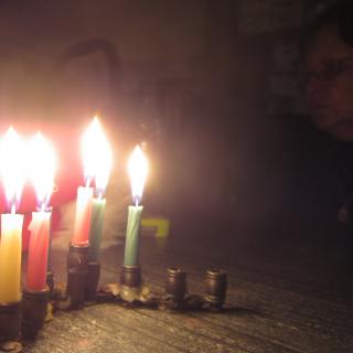 A small menorah with six lit candles, with a person's face obscured in the dark background