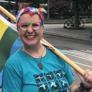 Helen Rose smiles, carrying the post of a rainbow flag over her shoulder. 