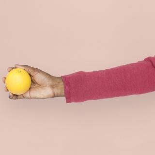 An outstretched arm, with dark skin, holds an orange.