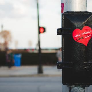 With a street and sidewalk blurred in the background, a red heart sticker is stuck to a streetpost. It says, "Make America Love Again."