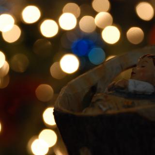 With blurred multicolored Christmas lights in the background, a small Jesus-in-the-manger from a nativity scene is in half-light.