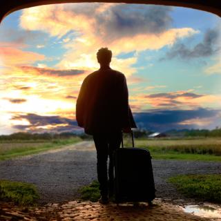 Through a large doorway, a person in silhouette rolls a suitcase into a road and a sky full of clouds.