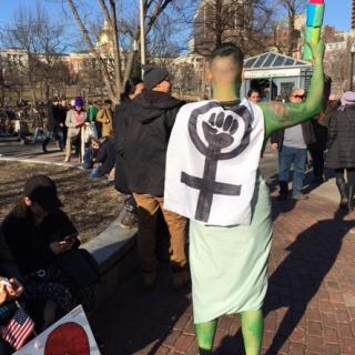 A young woman costumed as the Statue of Liberty with a cardboard torch stands in Boston Common, wearing a sign on her back that shows a woman symbol with a fist clenched in the circle at the top.