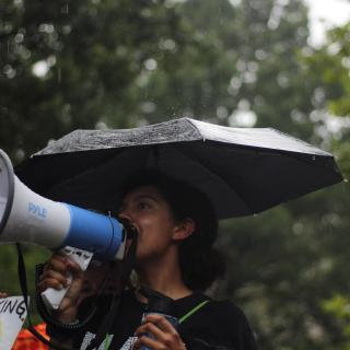 A person, smiling, speaks into a megaphone under an umbrella, with fellow protesters visible around her.