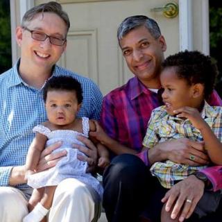 A family of two dads, a toddler son, and a baby daughter sit on a porch together.