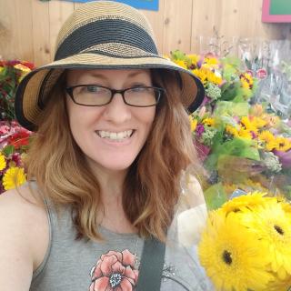 Monica Dobbins in a straw hat, holding flowers, with racks of sunflowers behind her.
