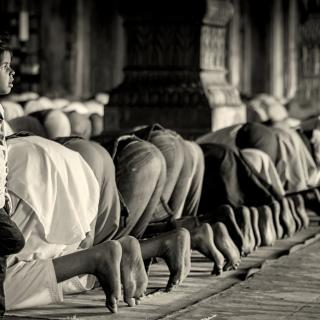 A child, lost in thought, leans against a pillar next to a row of Muslims bowing in prayer in a mosque
