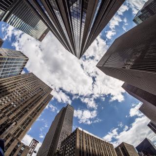 A view, straight upwards, of the skyscrapers around Rockefeller Center. In the negative space between buildings, a blue sky with clouds is visible.