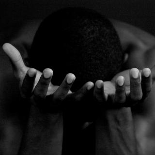An African-American person bows their head, bare shoulders visible, holding both hands open with palms up.