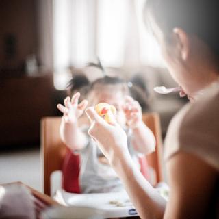 A parent, holding food on a spoon, in front of a toddler reaching.