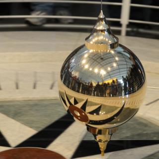 A gold Foucault's pendulum slowly swings across a marbled floor. 