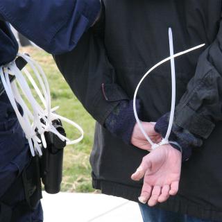 A person's hands are bound together behind their back with plastic zip-ties, and a police officer's holster, with more zip-ties, is in the frame.
