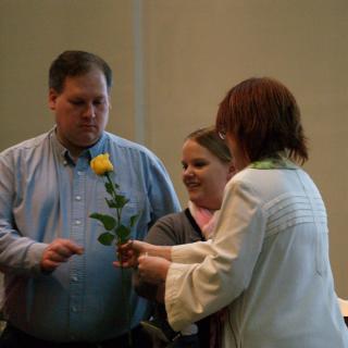 A minister handing a yellow rose to a church member, the lit chalice in the background