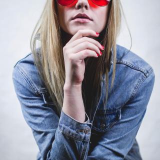 A young woman faces the camera, hand cupping her face, wearing glasses with bright red lenses. 