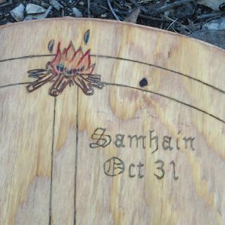 An arc of a wooden wheel, with "Samhain Oct 31" carved into it, along with a bonfire.