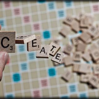 In the background, letters are piled on a Scrabble board. In the foreground, someone holds the "C" tile as the letters "R E A T E" fall towards the board.