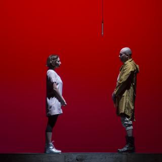 Against a red background, two people stand facing one another, with a gap between them.