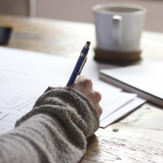 Person in sweater with hot beverage writing with a pen