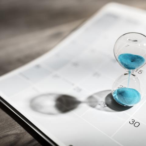 A clear hourglass filled with blue sand rests on the page of a calendar 