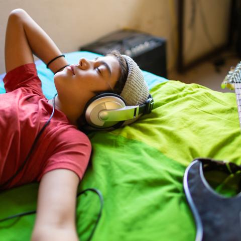 A young teen lies on their bed, eyes closed, listening to music on headphones. A guitar lies on the bed next to them.